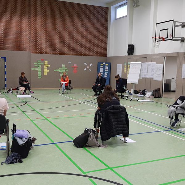 Teilnehmende sitzen in einer Sporthalle im Stuhlkreis und an der Wand hängen Plakate