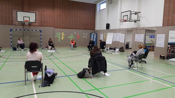 Teilnehmende sitzen in einer Sporthalle im Stuhlkreis und an der Wand hängen Plakate